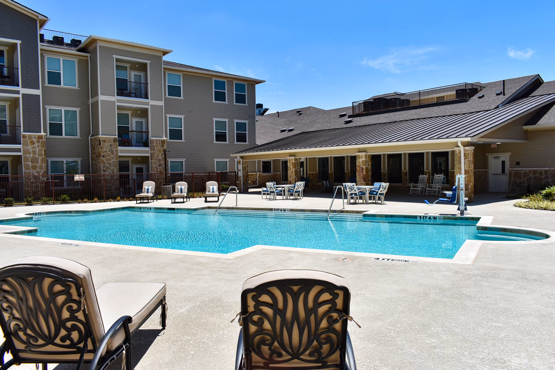 Outdoor swimming pool area at Legacy Oaks of Midlothian Senior Living with lounge chairs and tables around the pool, adjacent to a multi-story residential building under a clear blue sky.