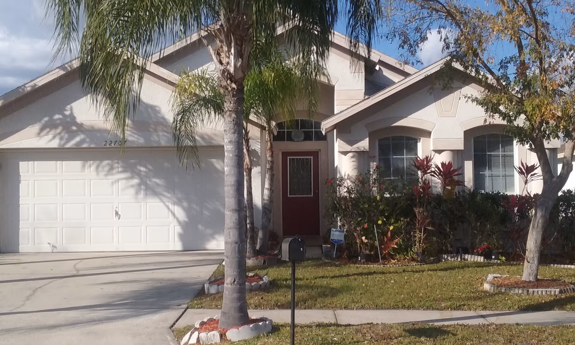 Front exterior view of a single-story house with a white garage door, a red front door with decorative glass, palm trees, and landscaped bushes and grass in the yard.