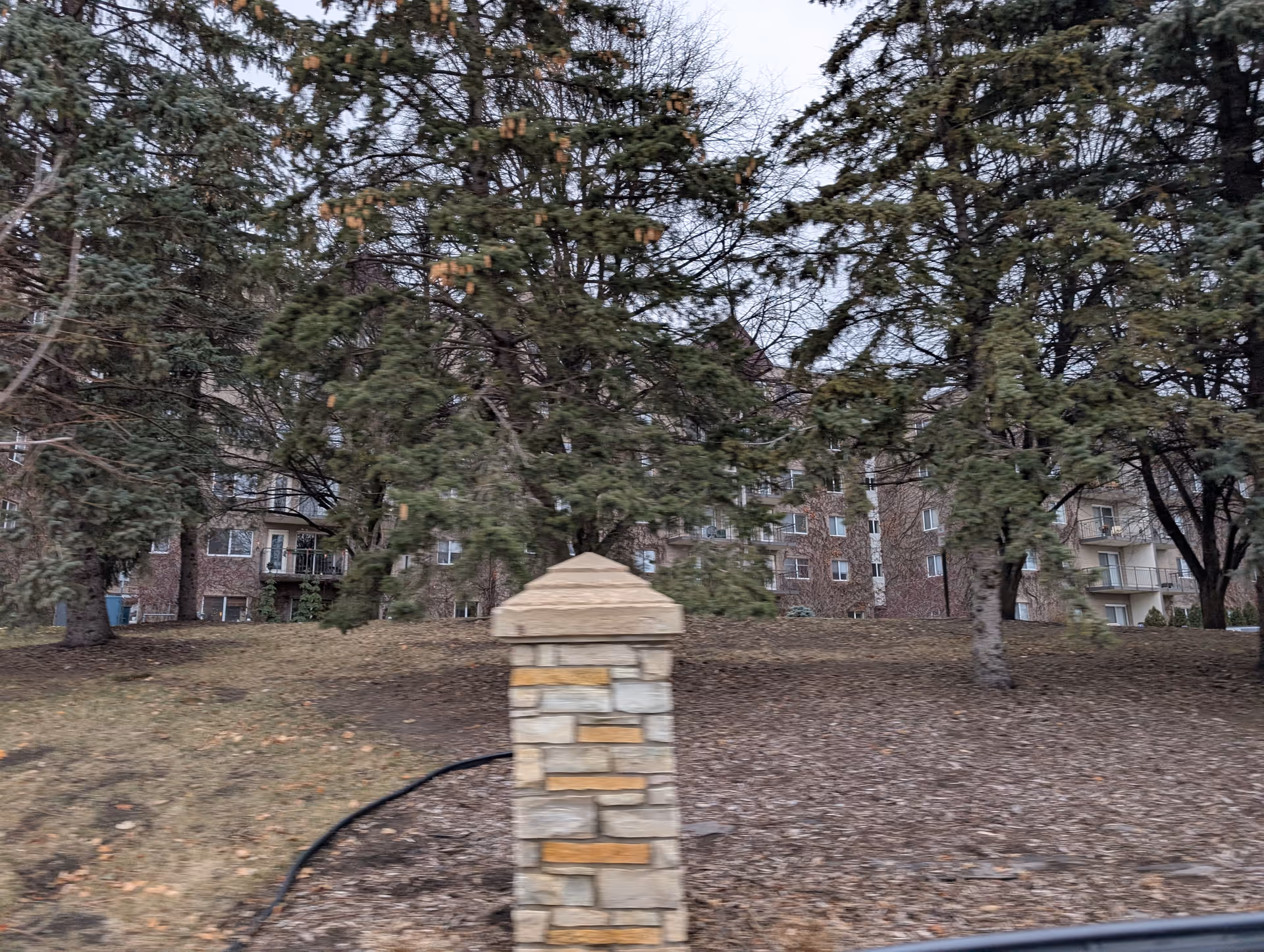 Stone post and grassy slope with evergreen trees partially obscuring a multi-story brick residential building.