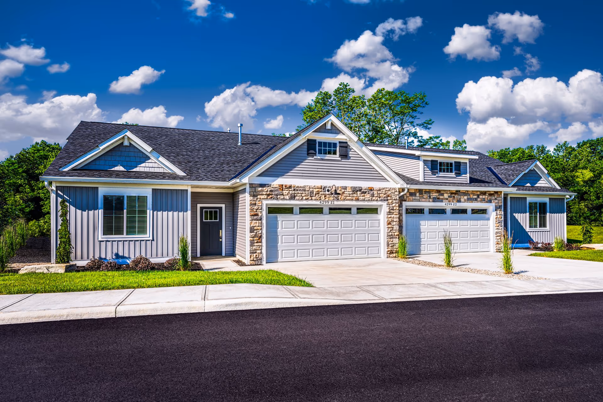 Exterior view of a modern residential building with two attached garages, stone and siding facade, and a well-maintained lawn under a blue sky with scattered clouds.