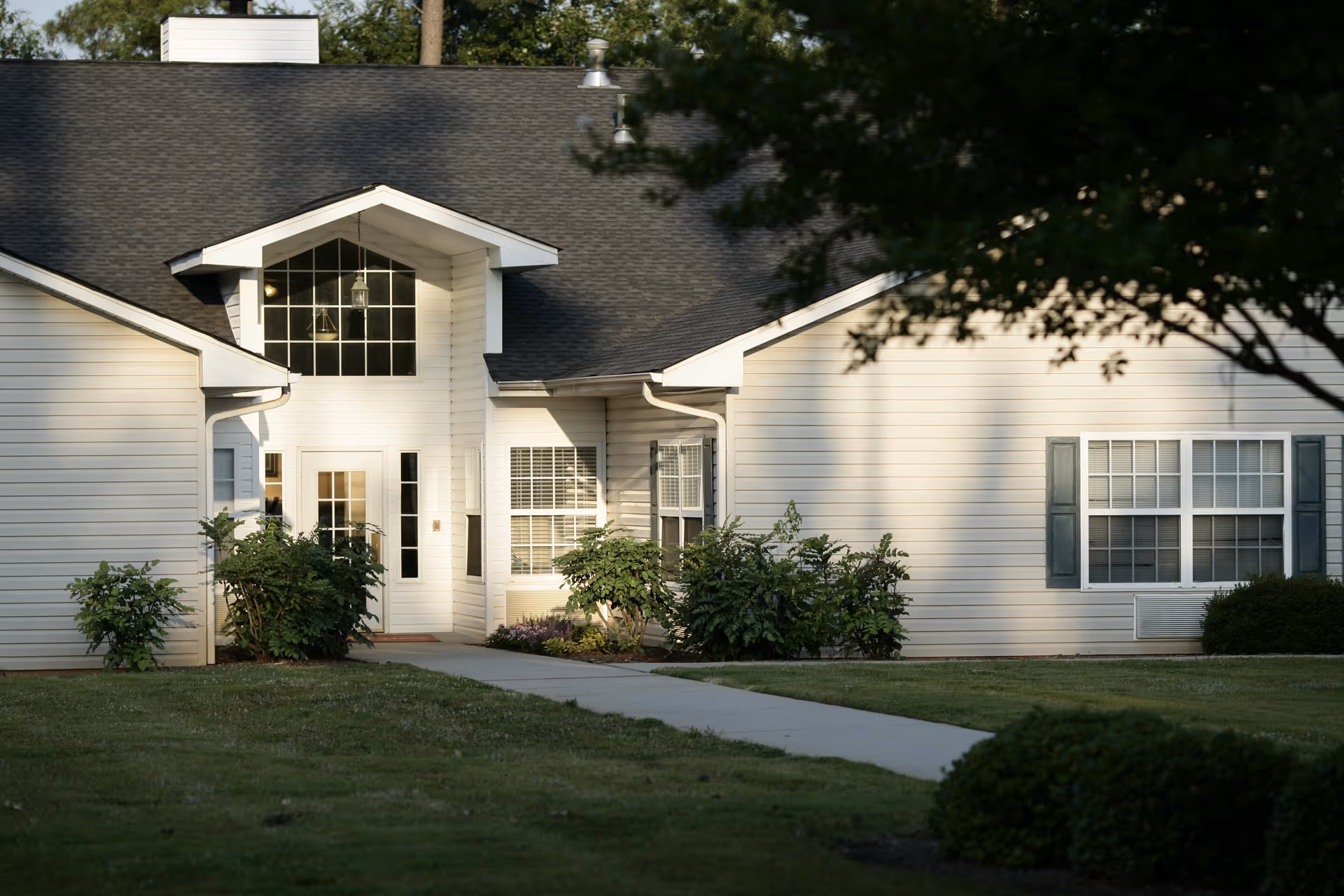 Exterior view of a single-story white building with a dark shingled roof, featuring a central entrance with a glass door and large windows above. The building is surrounded by green bushes and a well-maintained lawn with a concrete walkway leading to the entrance.