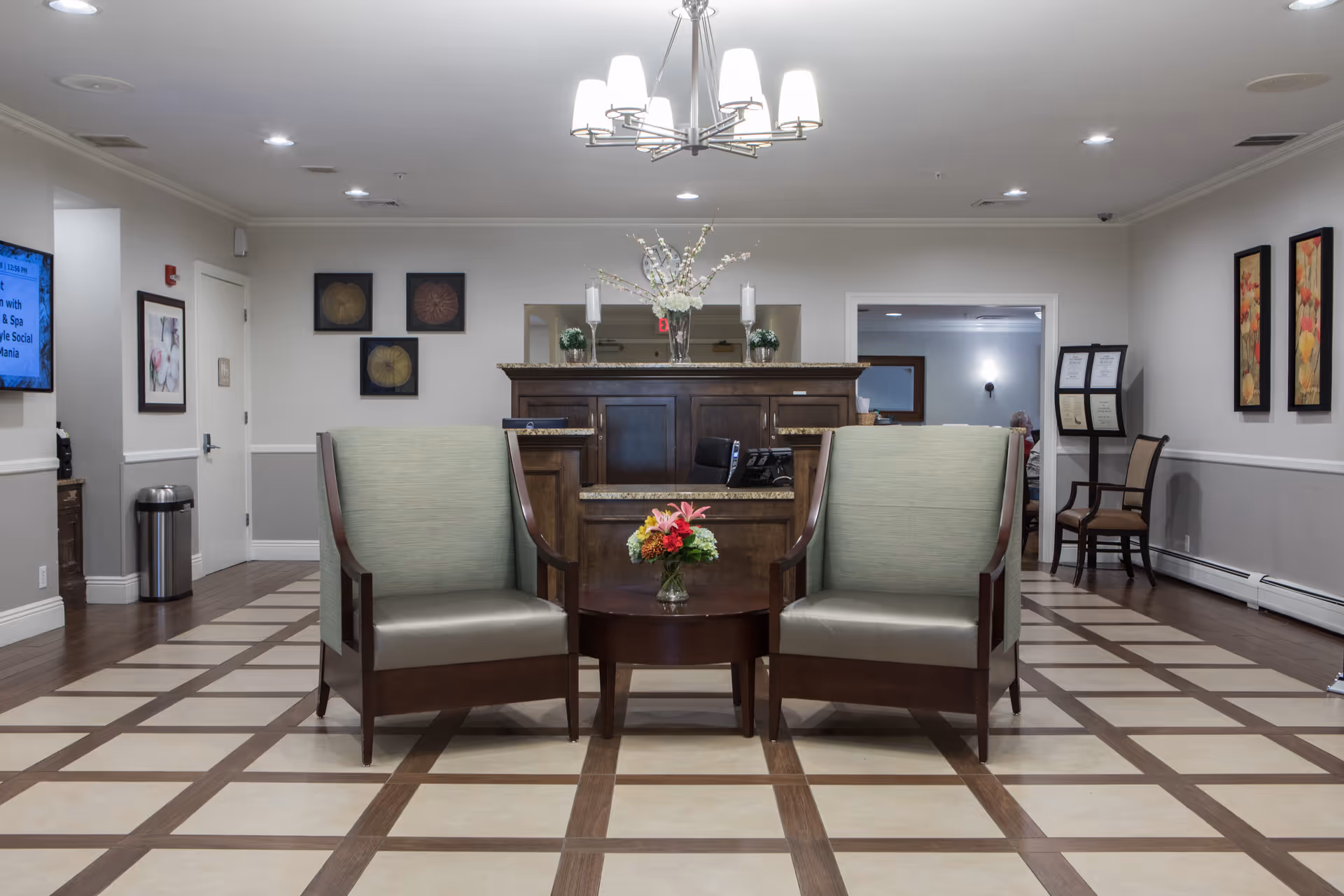 A spacious lobby with two green armchairs facing a wooden reception desk beneath a chandelier.