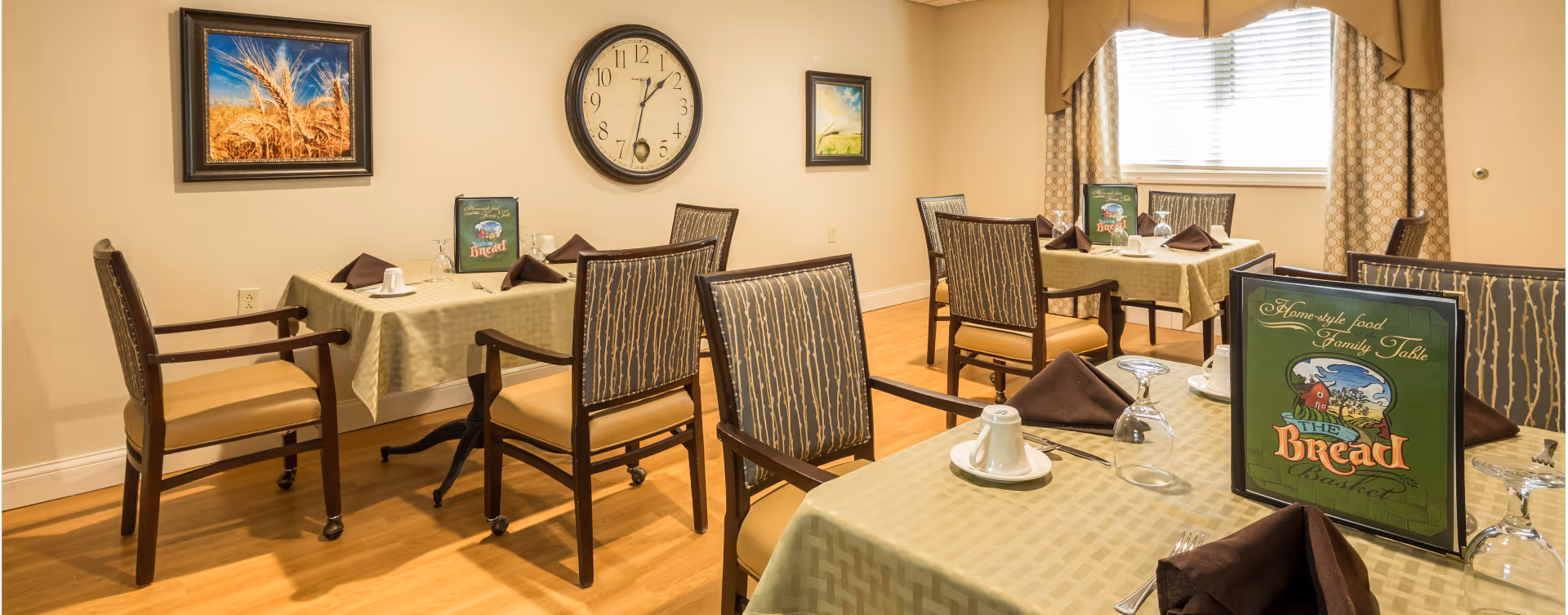 A dining room with several tables covered in light green tablecloths, each set with brown napkins, white cups, and upside-down wine glasses. The chairs have wooden frames with patterned upholstery. On the walls, there are two framed pictures and a large round clock. A menu titled 'The Bread Basket' is placed on each table. The room has wooden flooring and beige walls with a window covered by patterned curtains.