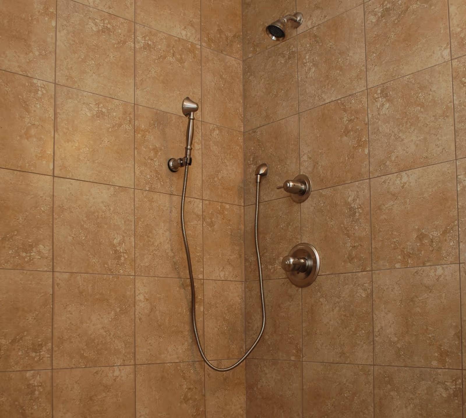A tiled shower area with beige stone tiles covering the walls. The shower features a fixed showerhead at the top and a handheld showerhead attached to a flexible hose, both with metallic finishes. There are two control knobs mounted on the wall for water temperature and flow.