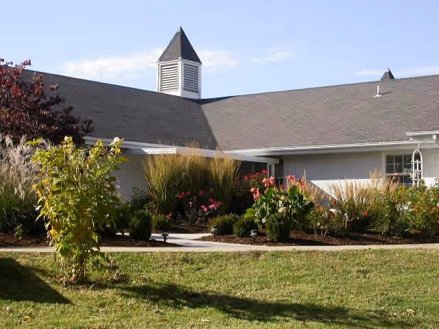 Exterior view of a single-story building with a gray roof and a small cupola on top. The building is surrounded by a well-maintained garden with various shrubs, flowering plants, and a green lawn under a clear sky.