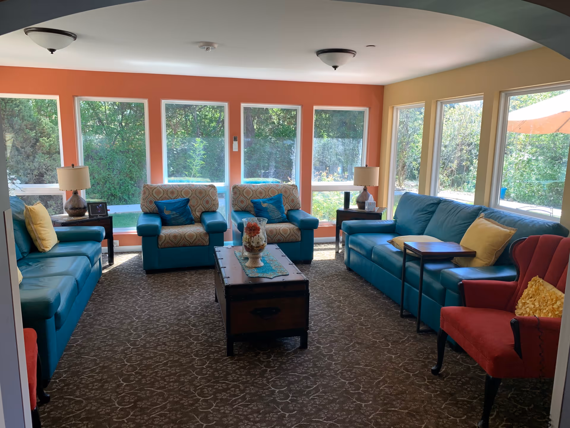 A bright living room with large windows showing greenery outside. The room has teal sofas and armchairs with patterned cushions, a red armchair with a yellow pillow, two side tables with lamps, and a wooden coffee table with a decorative centerpiece on a patterned carpet.