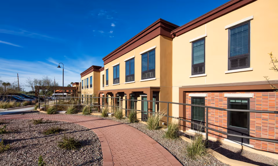 Exterior front of a two-story senior living building with a curved brick walkway, landscaping, and metal railing under a clear blue sky.