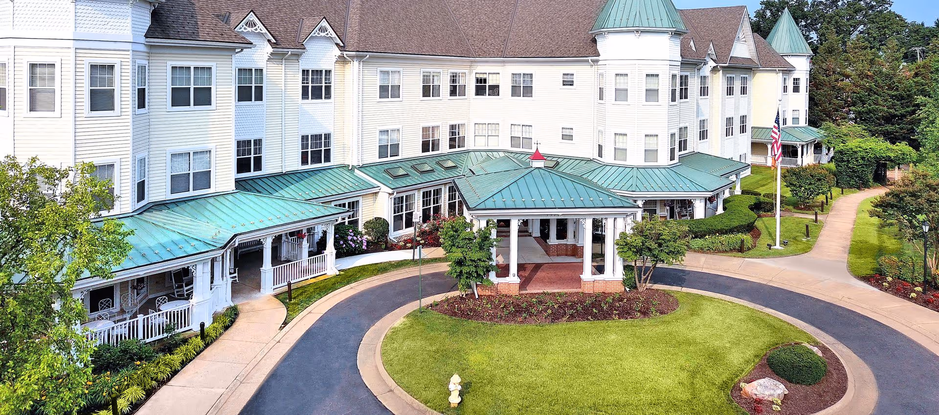 Front exterior of a multi-story senior living facility with a covered porte-cochere, circular driveway, green metal roofs and landscaped grounds.