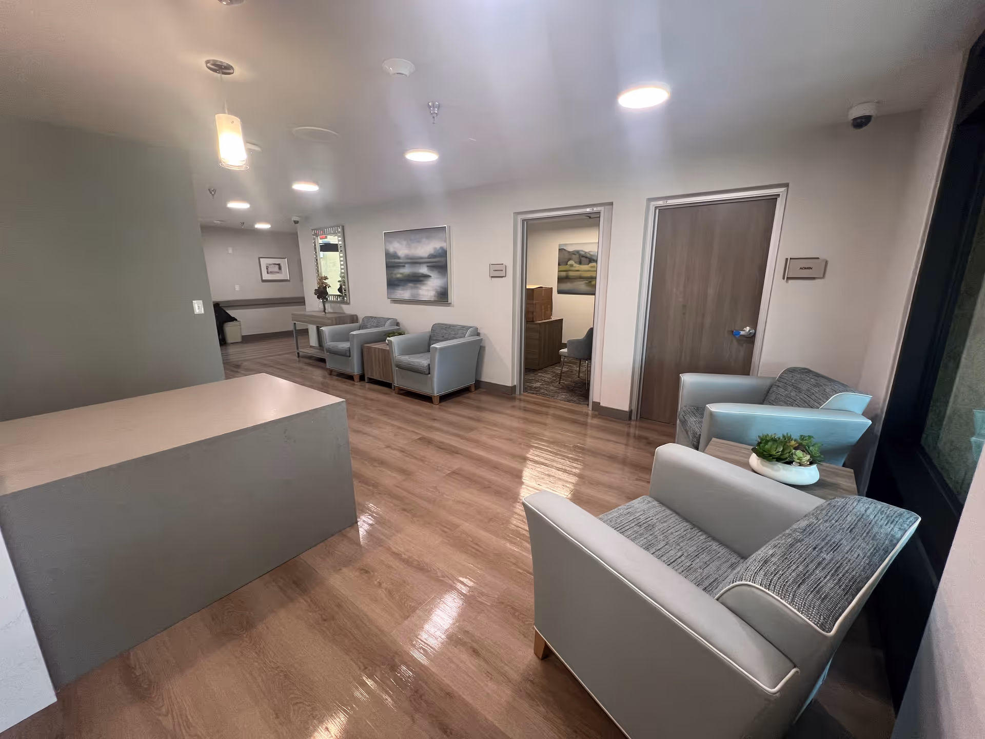 Modern senior living lounge with blue-gray chairs, wood floor, and doorway to private rooms.