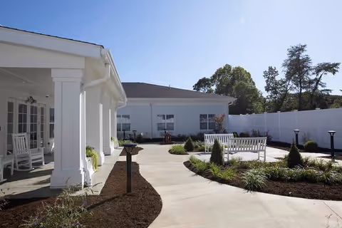 Outdoor courtyard area of a senior living facility with white benches, landscaped garden beds, a concrete walkway, and a white fence surrounding the space. The building exterior is white with large windows and a covered porch with rocking chairs.