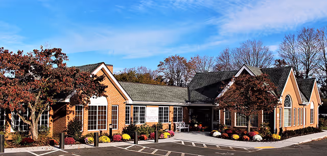 Brick single-story senior living facility building with a landscaped entrance, flowering shrubs, and a clear blue sky.