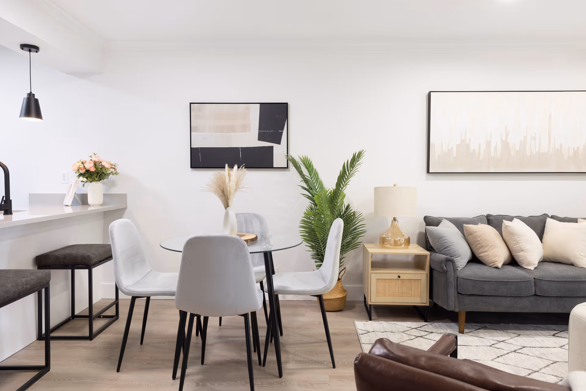A modern living and dining area featuring a round glass dining table with four gray upholstered chairs, a gray sofa with multiple cushions, a wooden side table with a lamp, a potted plant, and abstract wall art. The space has light wood flooring and a white wall with a kitchen counter and two bar stools on the left side.