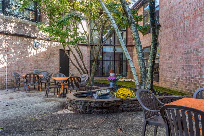 Outdoor courtyard area with brick walls surrounding it, featuring several plastic chairs and tables with orange tablecloths. There is a circular stone planter with trees and colorful flowers in the center, and large windows on the building walls.