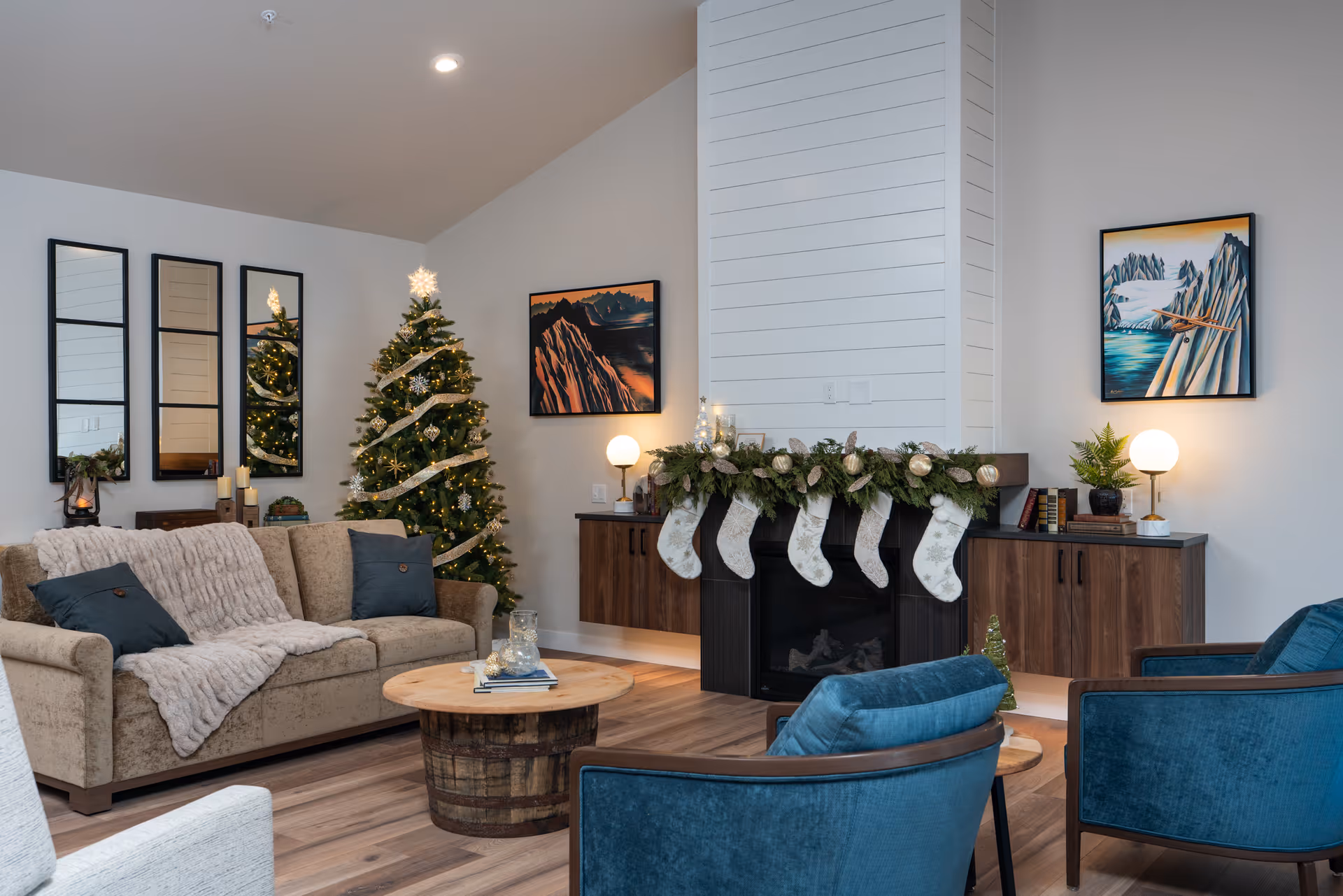 A cozy living room decorated for Christmas with a beige sofa draped with a light gray throw blanket and dark blue pillows, two blue armchairs, a round wooden coffee table, a decorated Christmas tree with lights and ribbon, a fireplace adorned with a garland and five white stockings, wooden cabinets on either side of the fireplace with table lamps, and two framed landscape paintings on the walls.