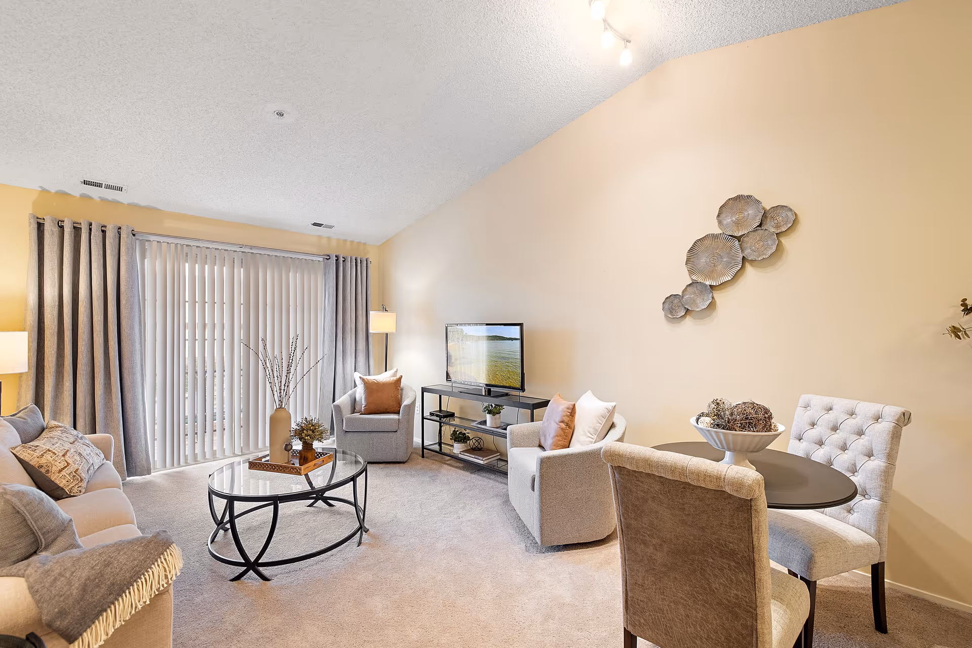 A bright living room with beige walls and carpet, featuring a beige sofa with cushions, two armchairs, a glass coffee table with decorative items, a small round dining table with four upholstered chairs, a TV on a black metal stand, and large vertical blinds covering a sliding glass door.