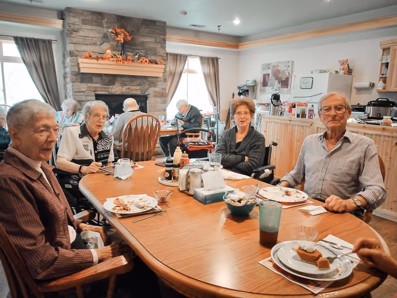 A group of elderly people sitting around a wooden dining table in a cozy room with a stone fireplace and windows with curtains. They are eating and socializing, with plates of food and drinks on the table.