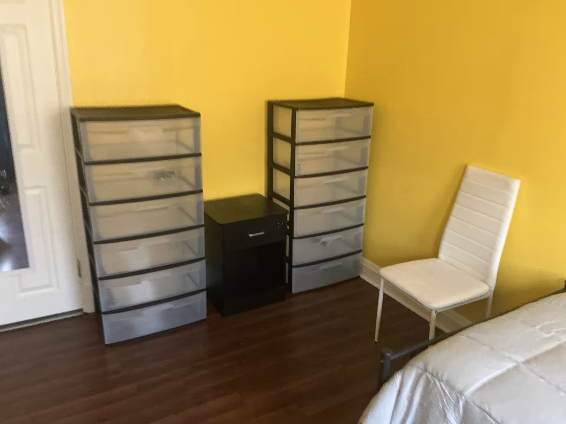 Yellow-walled bedroom with plastic drawer storage units, a white chair, and a bed.