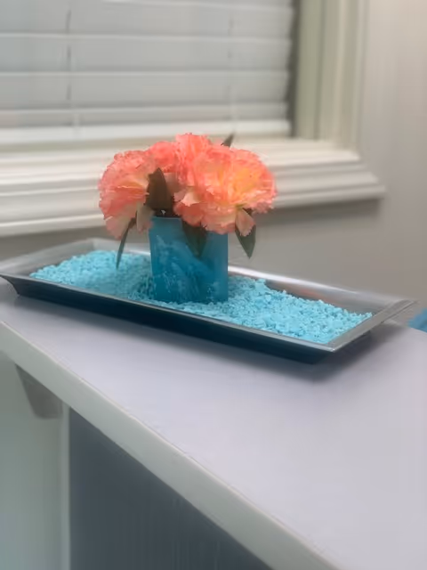 Pink carnations in a blue vase on a turquoise bead tray by a window.