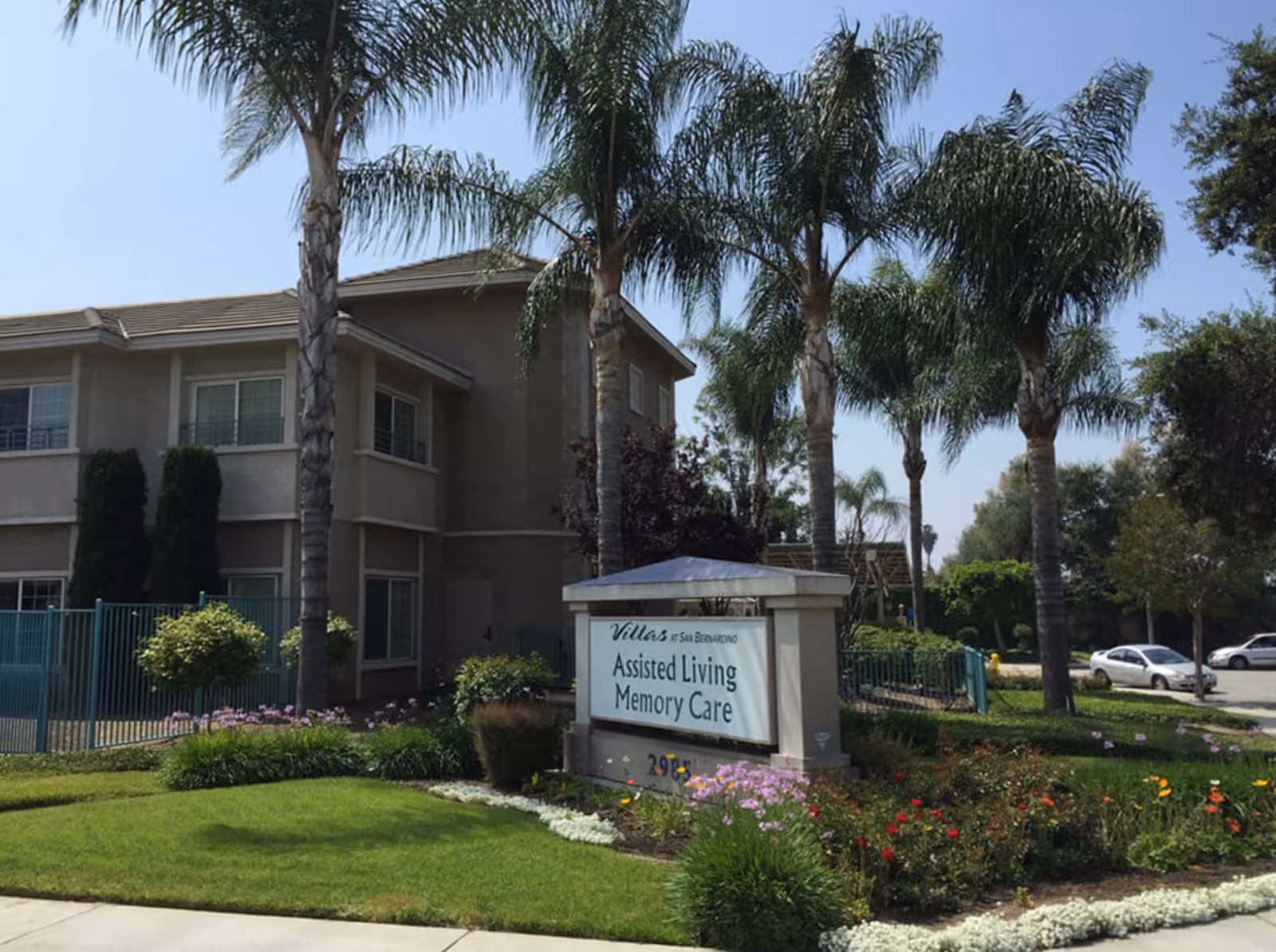 Exterior view of Villas at San Bernardino assisted living and memory care facility with palm trees, landscaped garden, and a sign displaying the facility name.