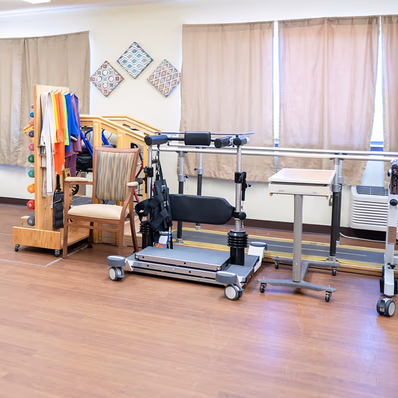 A physical therapy room with parallel bars for walking assistance, a chair, a rack holding colorful exercise bands and small dumbbells, and a small rolling table. The room has wood flooring and beige curtains covering the windows.