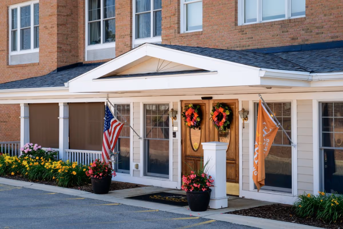 Front entrance of a senior living building with double wooden doors topped by a small portico, wreaths on the doors, an American flag and an orange flag, and potted flowers along the walkway.