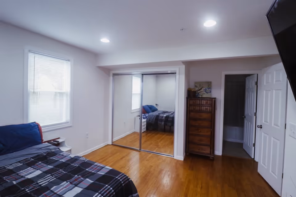 Bedroom in a senior living facility featuring a bed, dresser, window, and mirrored closet doors.