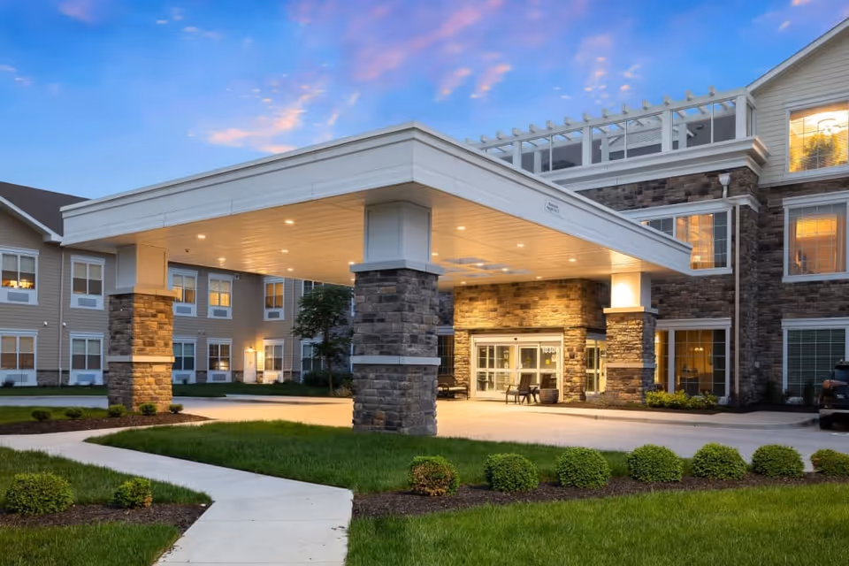Front entrance of a senior living building with a covered porte-cochere, stone columns, lit entryway, and landscaped lawn at dusk.