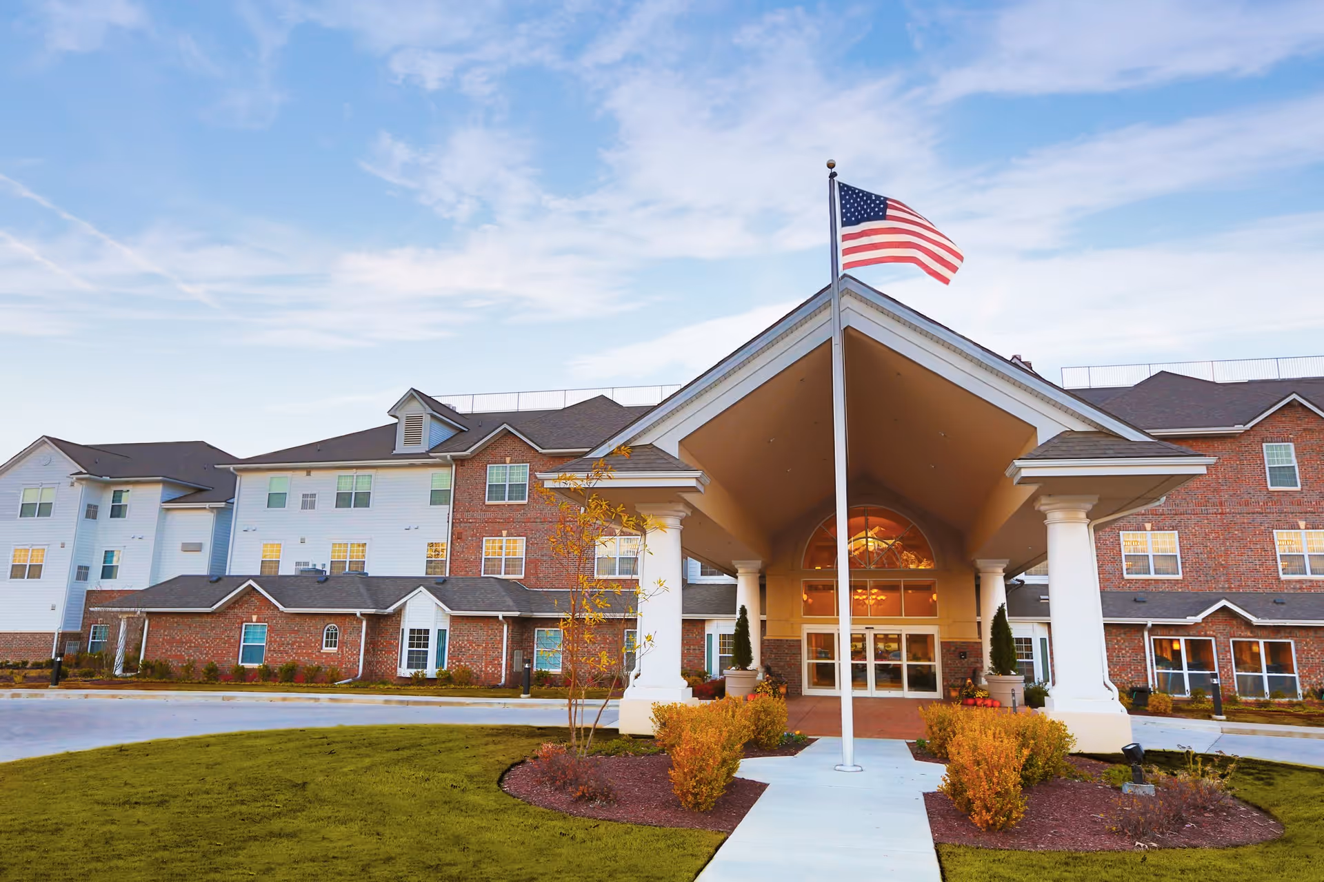 Front exterior view of Robinwood Retirement Resort, a large multi-story building with a covered entrance supported by white columns. An American flag flies on a flagpole in front of the entrance, and there are landscaped bushes and a lawn surrounding the walkway leading to the doors. The sky is partly cloudy with a soft evening light.