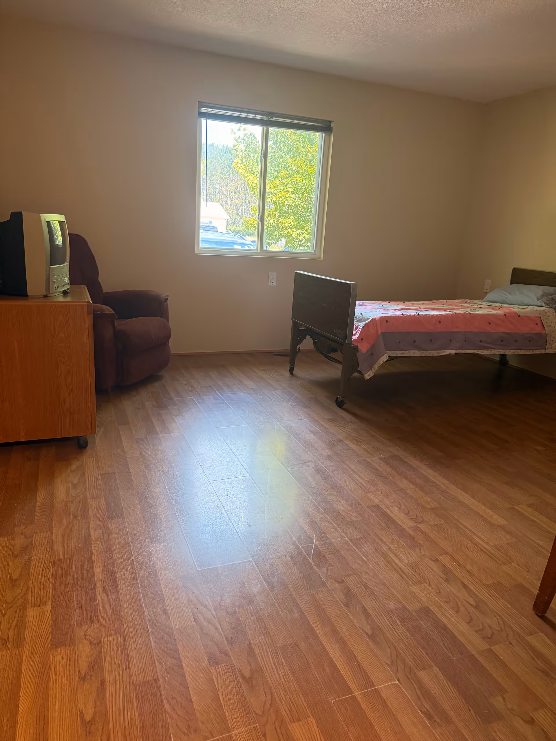 A sparsely furnished bedroom featuring a bed, window, recliner, and a TV on a dresser.