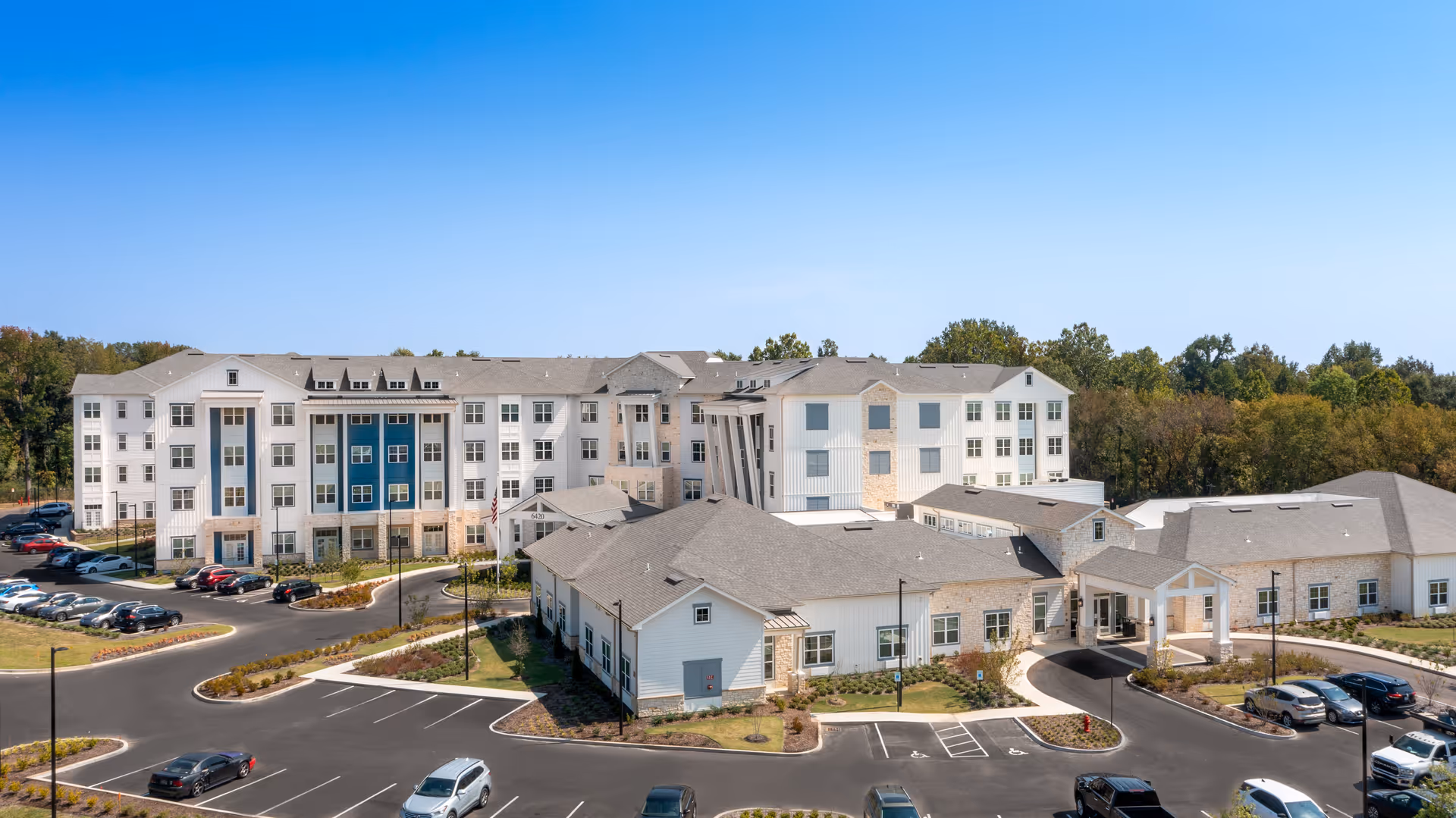 A large, modern senior living facility building with multiple floors and a combination of white and blue exterior walls. The building is surrounded by a parking lot with several cars parked and landscaped greenery. Trees and a clear blue sky are visible in the background.