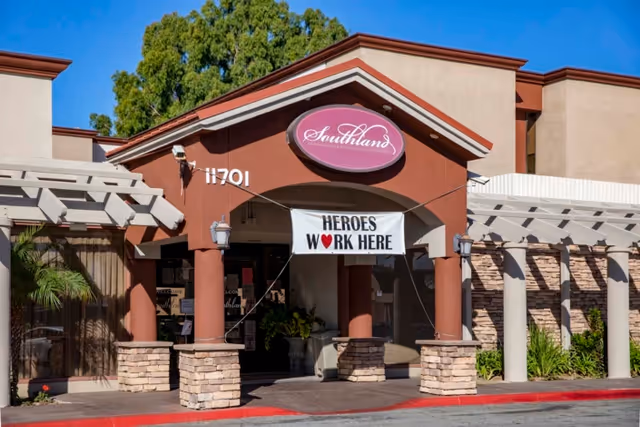 Entrance of Southland Care Center building with a sign above the door reading 'Southland' and a banner hanging below that says 'HEROES WORK HERE' with a red heart symbol. The building has a brown and beige exterior with stone pillars and a pergola on the right side.