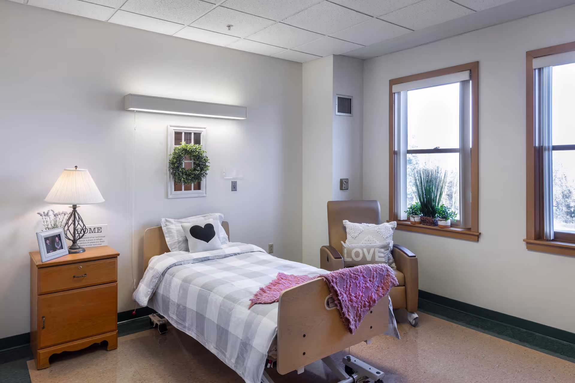 A cozy bedroom in a senior living facility featuring a single bed with a checkered blanket and heart-shaped pillow, a wooden nightstand with a lamp, framed photo, and decorative sign, a comfortable armchair with a pillow that says 'LOVE', and two windows with plants on the windowsill letting in natural light.
