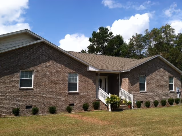 Exterior view of a single-story brick residential building with a gabled roof, white-framed windows, a small front porch with white railings, and a well-maintained lawn with small shrubs along the foundation. Trees and a partly cloudy blue sky are visible in the background.