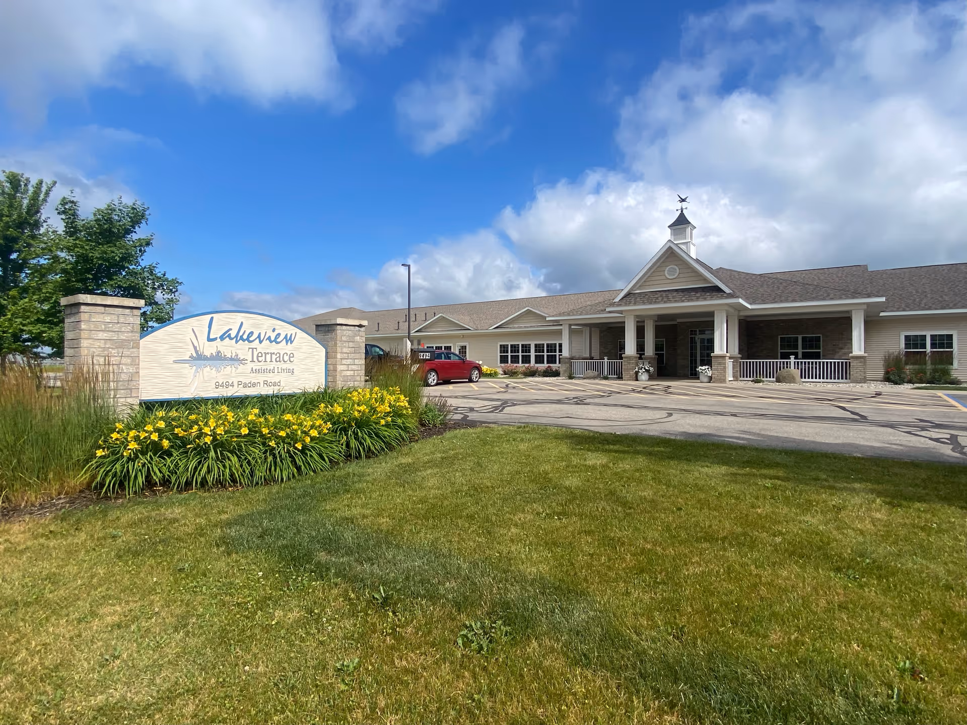 Exterior view of Lakeview Terrace Assisted Living facility on a sunny day with a partly cloudy blue sky. The building has a beige facade with a covered entrance and a weather vane on top. In front of the building is a parking area with a red car and a landscaped area with green grass, yellow flowers, and a sign that reads 'Lakeview Terrace Assisted Living 9494 Paden Road'.