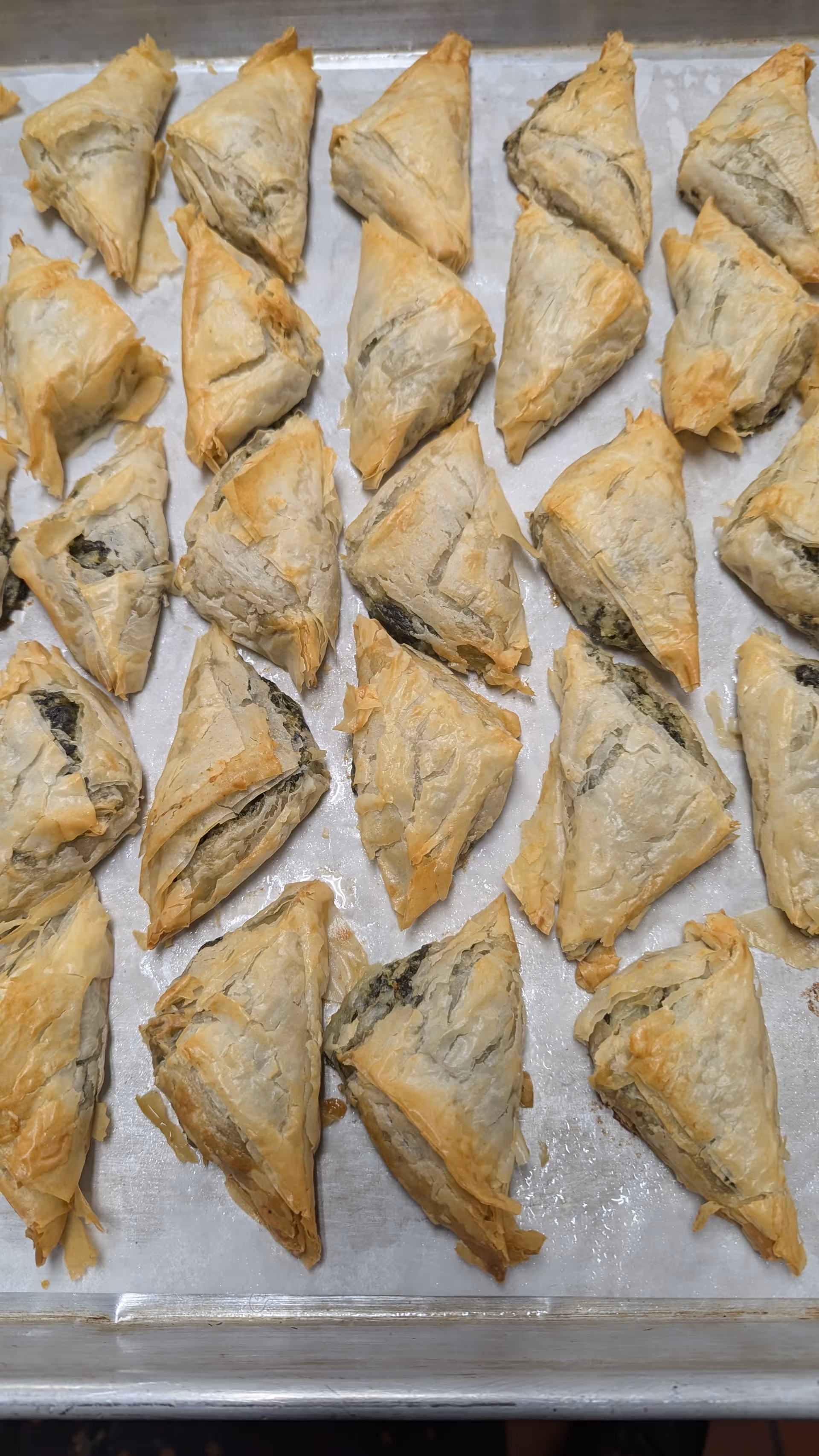 Rows of golden-brown triangular pastries arranged on a baking sheet in a kitchen.