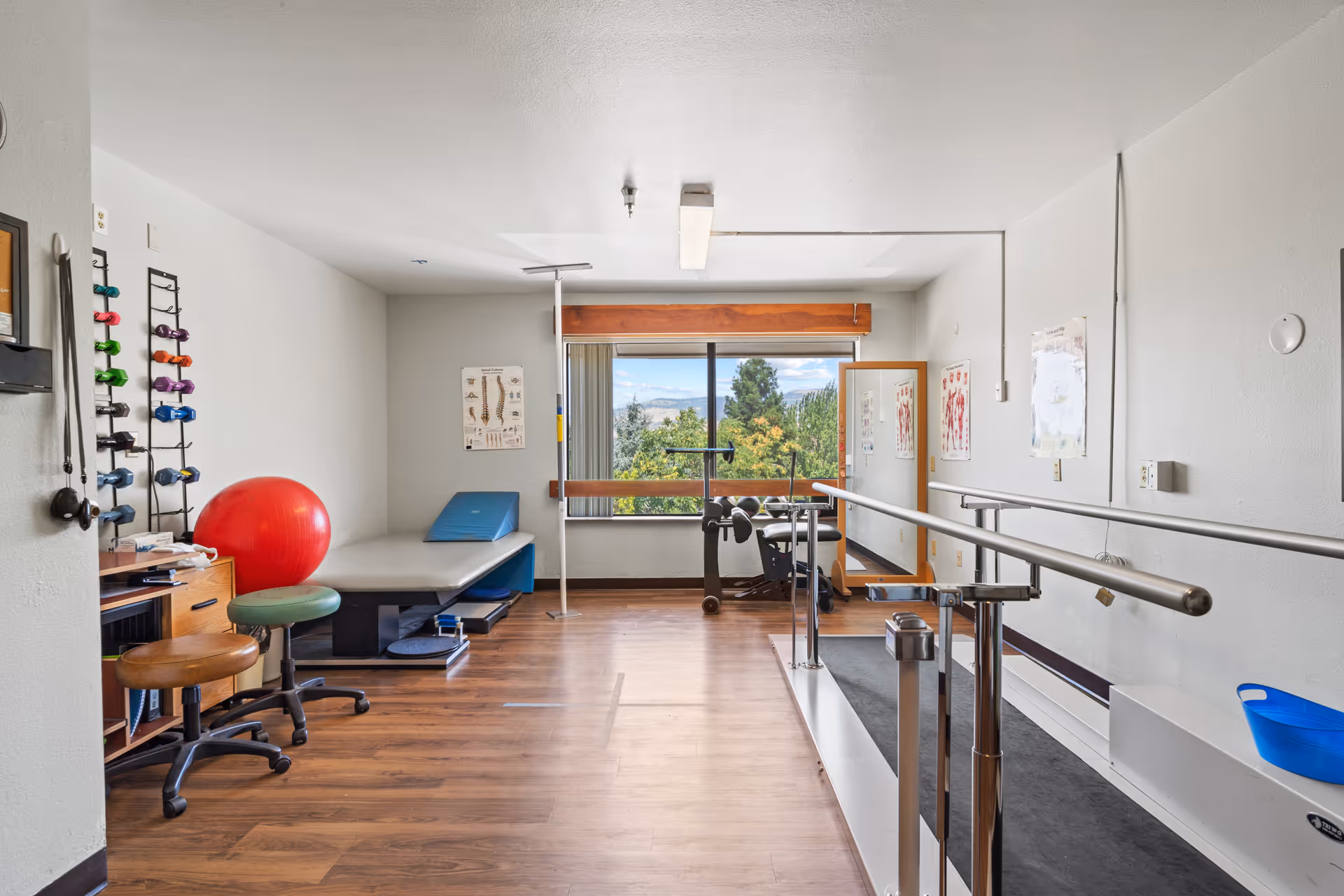 A rehab exercise room with parallel bars, therapy equipment, mirrors, and a window view.