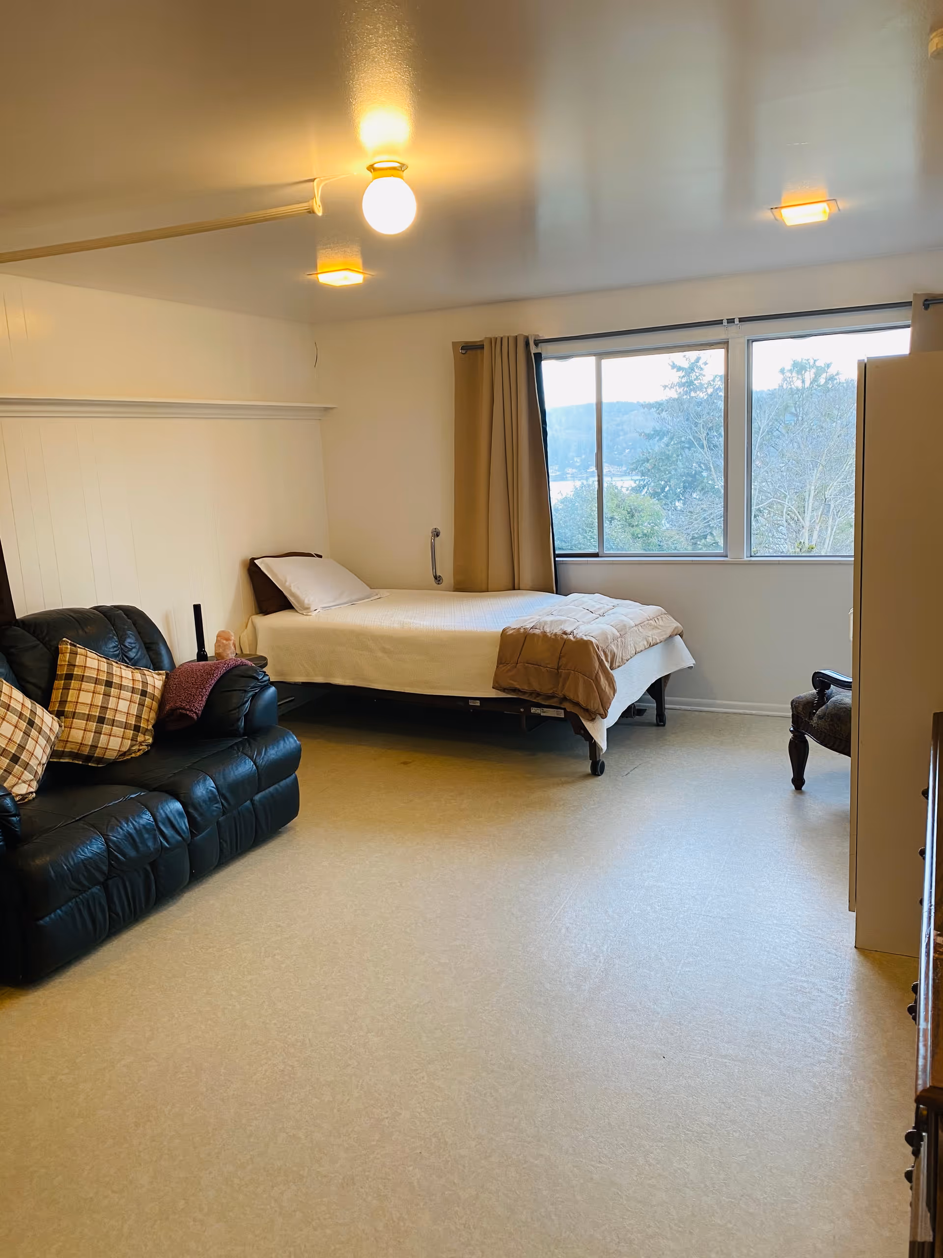 Bed and black leather sofa sit in a bright room with a large window.