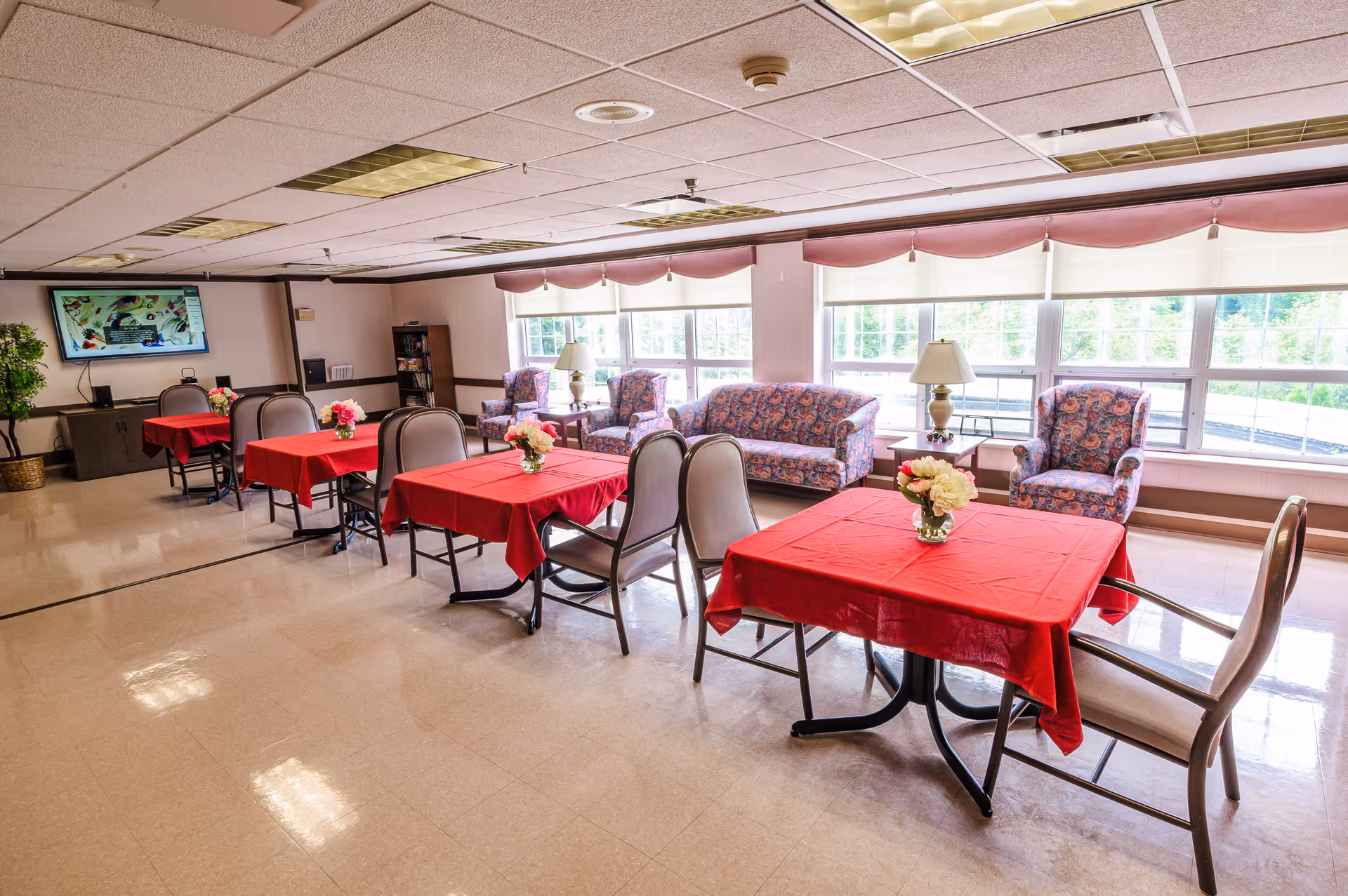 Senior living common area with red-draped dining tables and floral centerpieces.