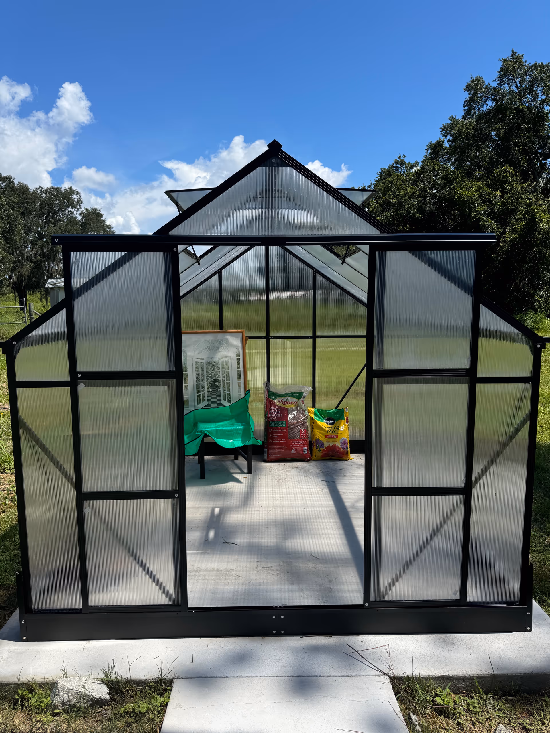 Glass-paneled greenhouse with bags and a chair inside, set in a grassy outdoor area.