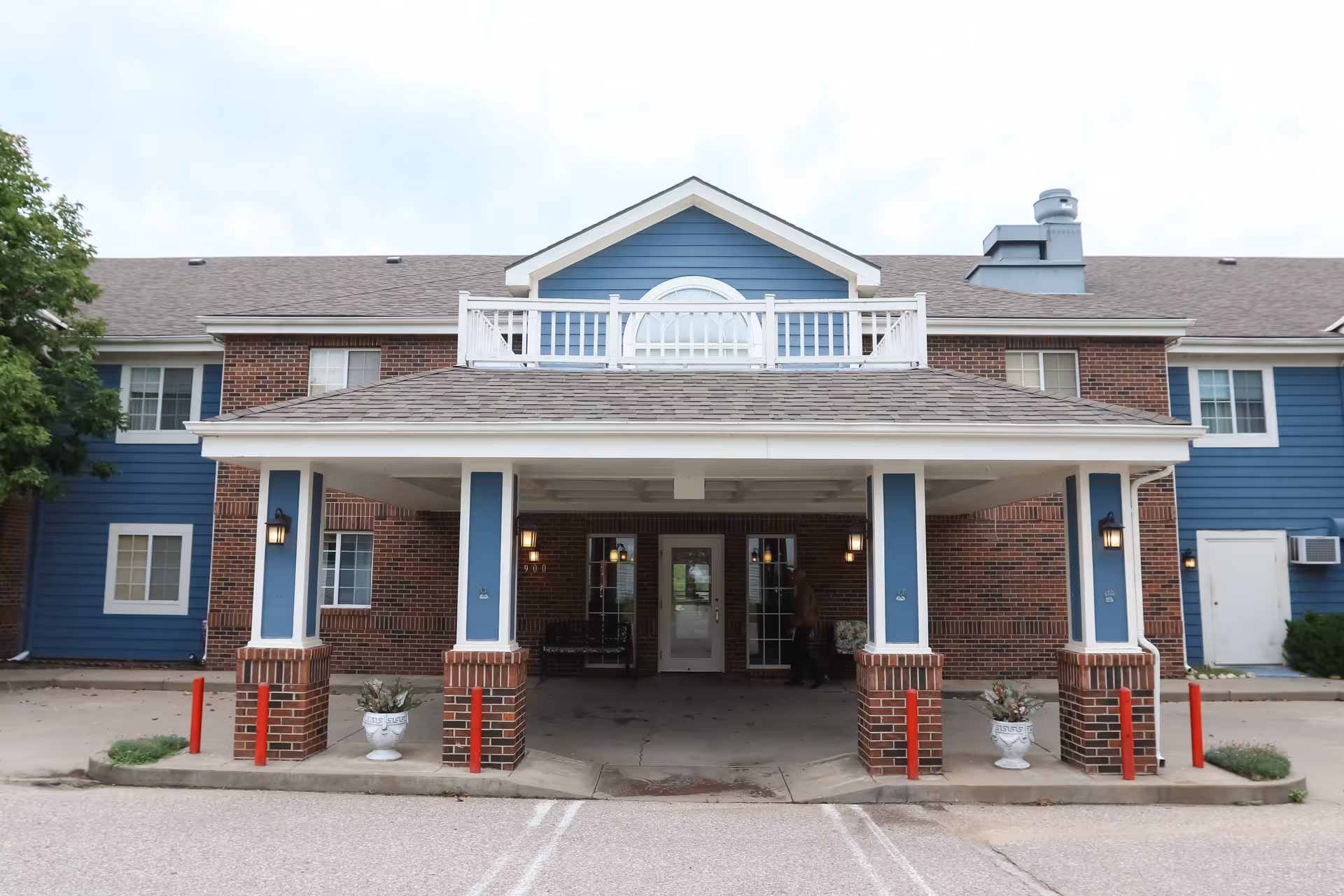 Front exterior view of a two-story building with blue siding and red brick accents. The entrance features a covered porch supported by four brick columns with blue panels. There are two white planters with plants on either side of the entrance. The building has multiple windows and a small balcony above the porch.