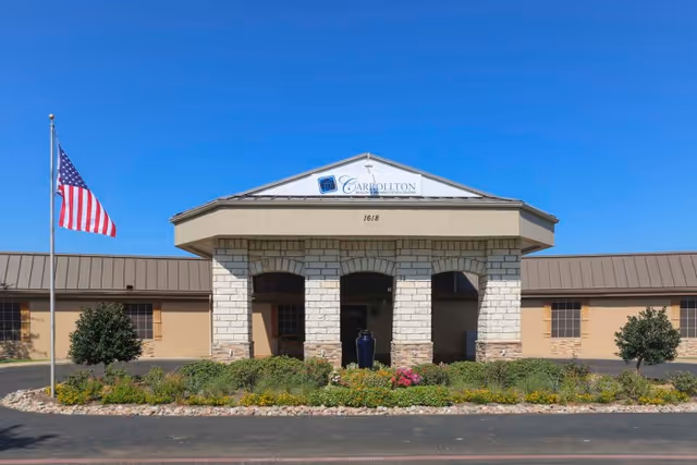 Front entrance of a single-story health and rehabilitation center with stone columns, landscaped flower beds, and an American flag.