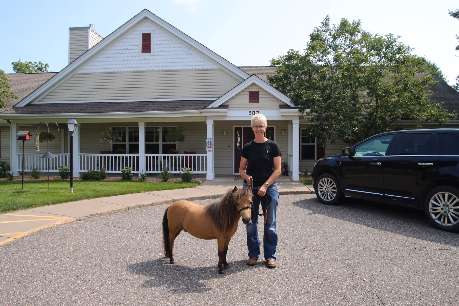 A person with short blonde hair wearing glasses, a black t-shirt, and blue jeans is standing outside a beige house with white trim, holding the leash of a small brown pony. There is a black SUV parked to the right, and the house has a porch with white railings and hanging flower baskets.