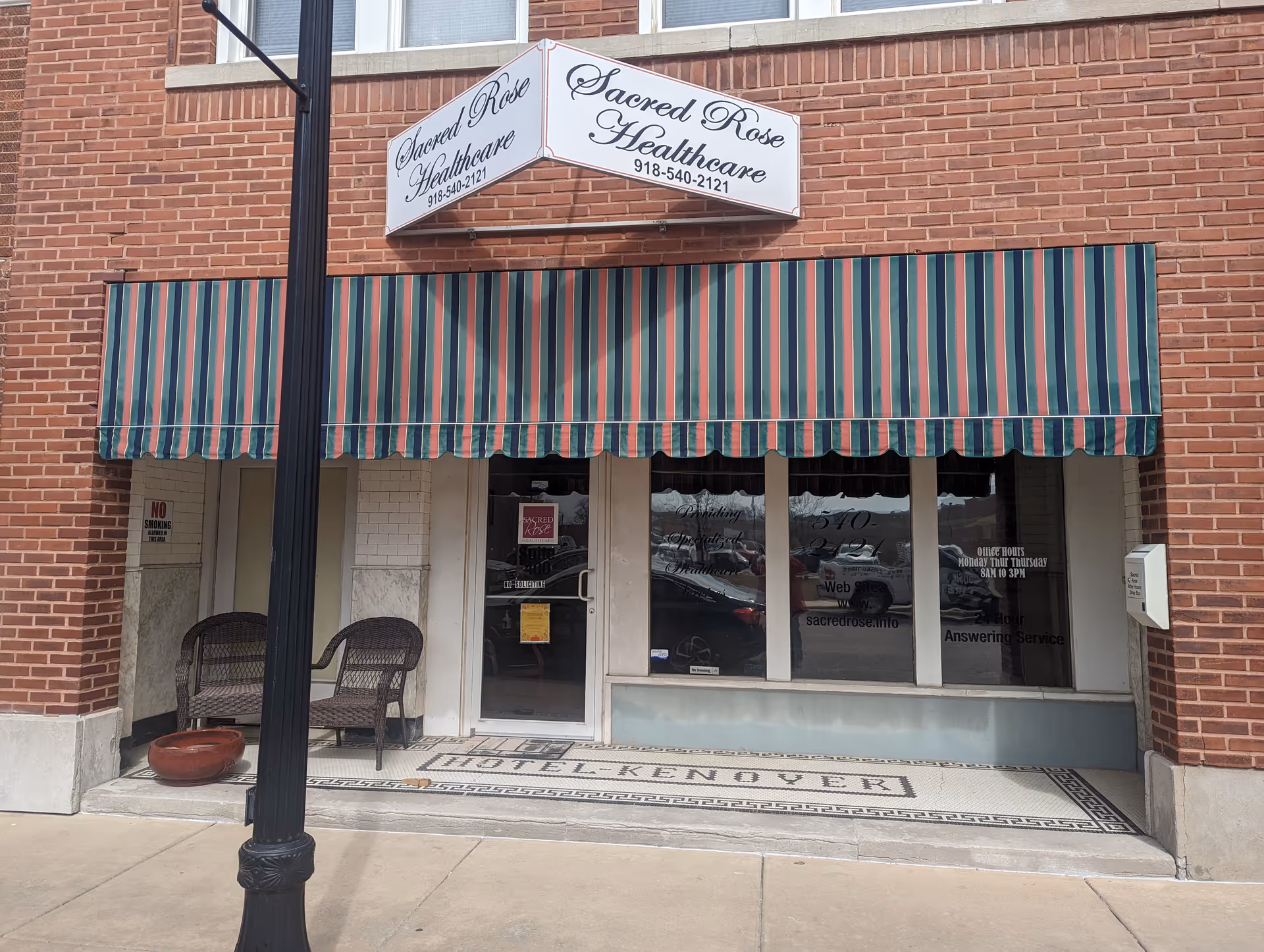 Brick storefront with striped awning, Sacred Rose Healthcare sign, glass door, two chairs outside entrance.