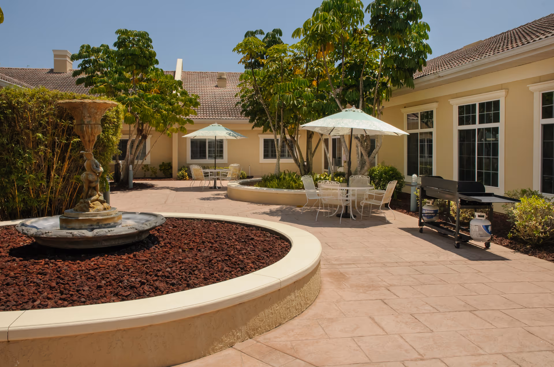 Outdoor courtyard area at Life Care Center of Port St. Lucie featuring a circular stone fountain, patio tables with umbrellas, chairs, trees, and a barbecue grill next to a beige building with multiple windows under a clear blue sky.