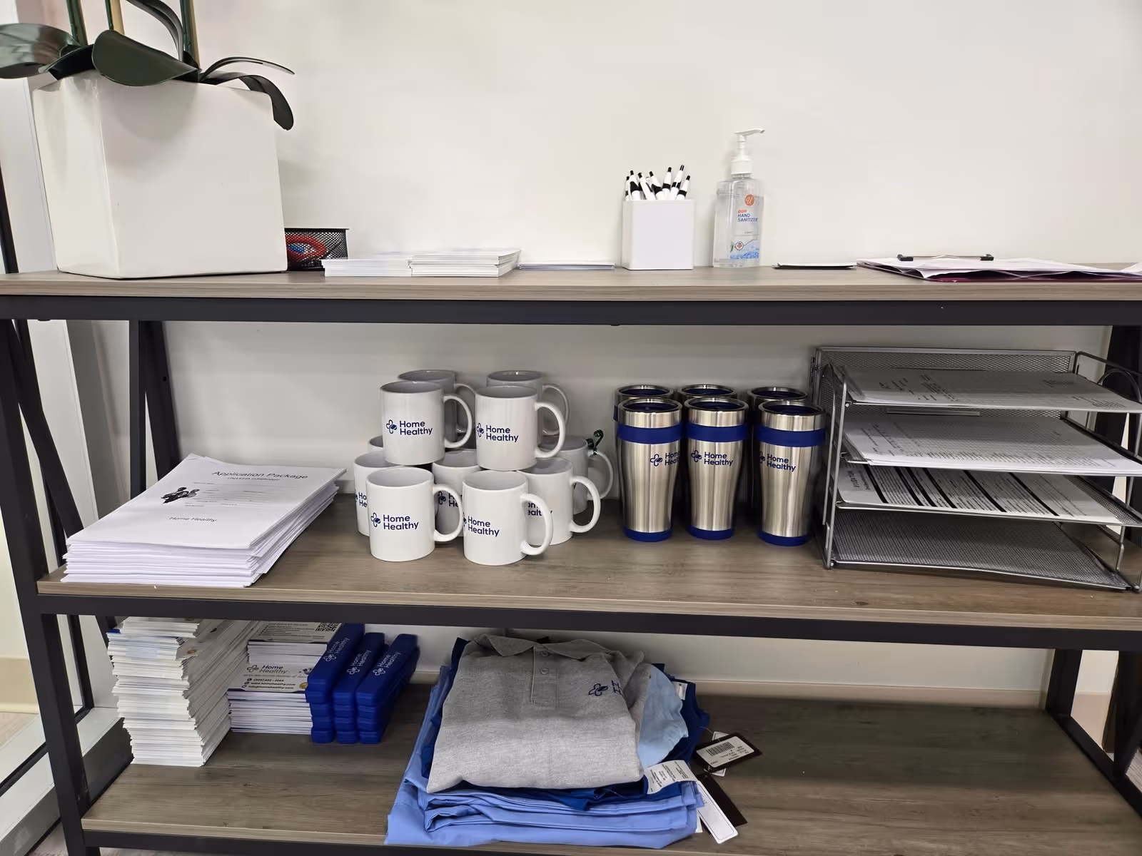 Shelf with Home Healthy mugs and tumblers, sanitizer, documents, and folded scrubs.