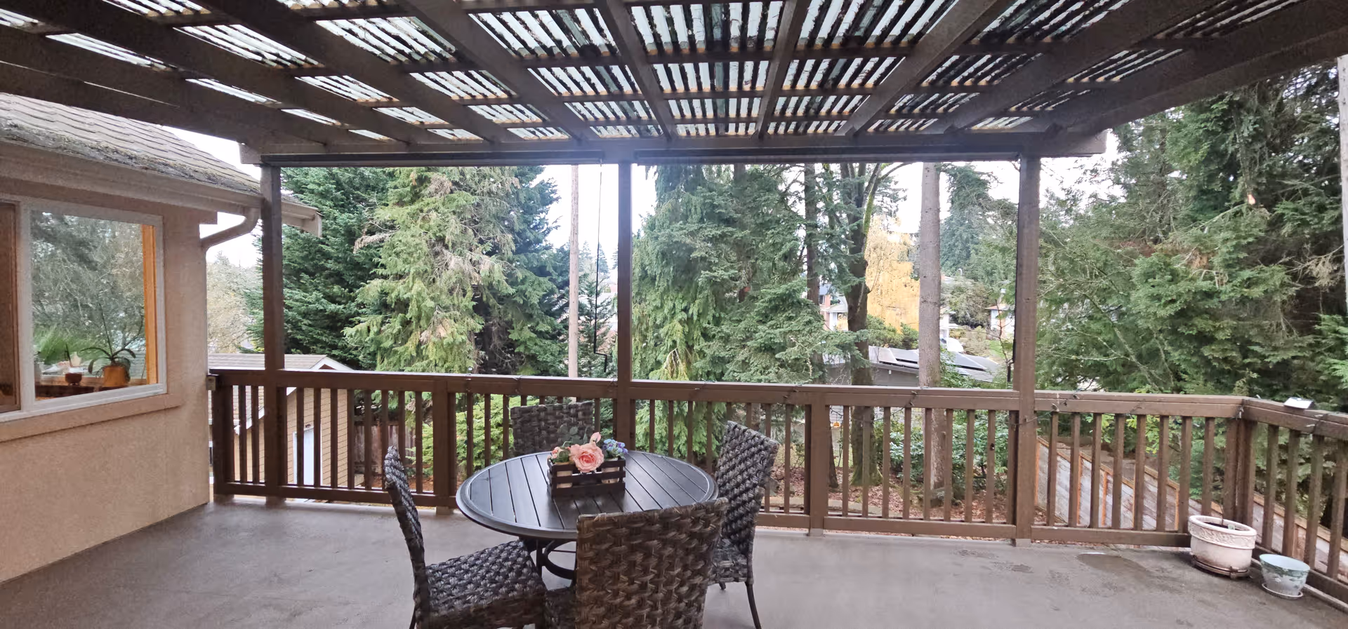 Covered outdoor balcony with a round table, four wicker chairs, and a view of trees.