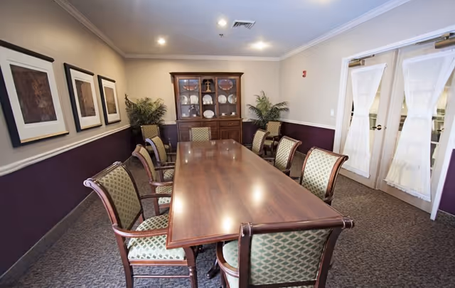 A formal dining room with a long wooden table surrounded by eight upholstered chairs. The walls are painted beige and purple with three framed artworks on one side. At the far end of the room, there is a wooden cabinet displaying plates and decorative items. Two potted plants are placed on either side of the cabinet. Double doors with white curtains are visible on the right side.