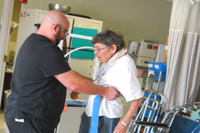 A caregiver assists an elderly woman with a mobility belt in a rehabilitation or care center room equipped with medical and mobility equipment such as walkers and wheelchairs.