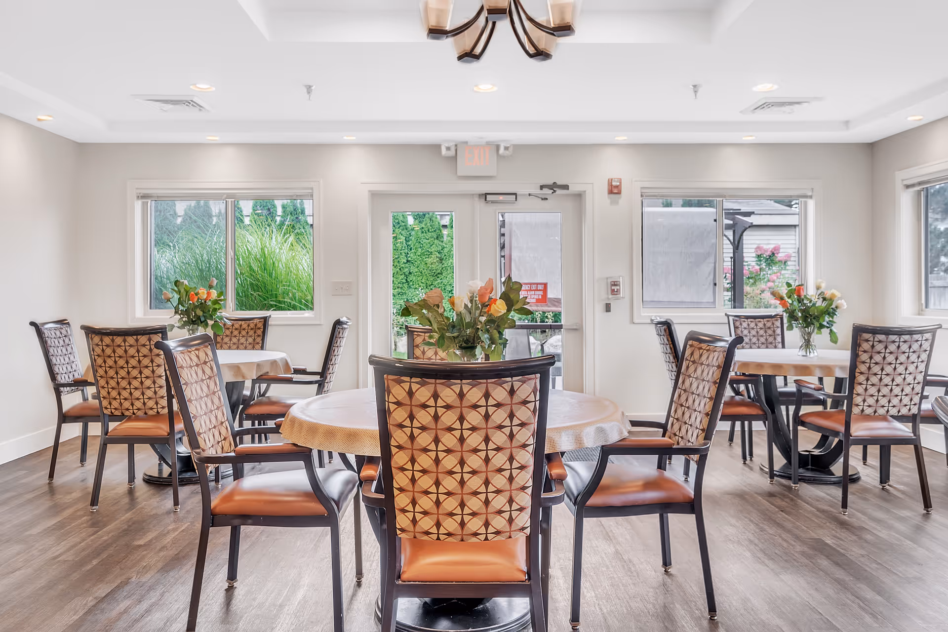 Bright dining room with round tables, patterned chairs, floral centerpieces, and windows showing greenery outside.