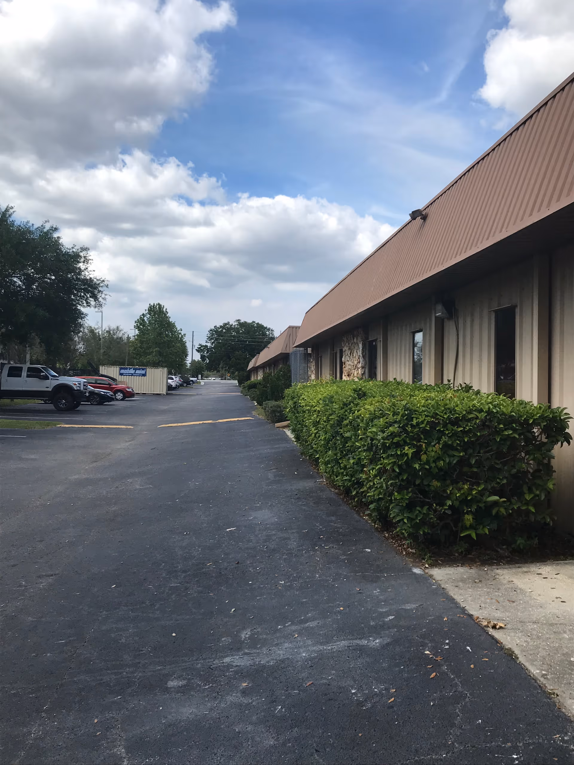 Long asphalt driveway beside a beige, single-story facility with trimmed hedges and parked cars.